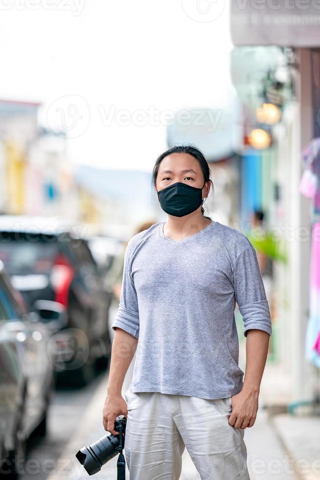 asiatischer professioneller kameramann trägt schwarze maske und hält spiegellose kamera, steht auf fußweg neben parkplatz in der stadt. foto