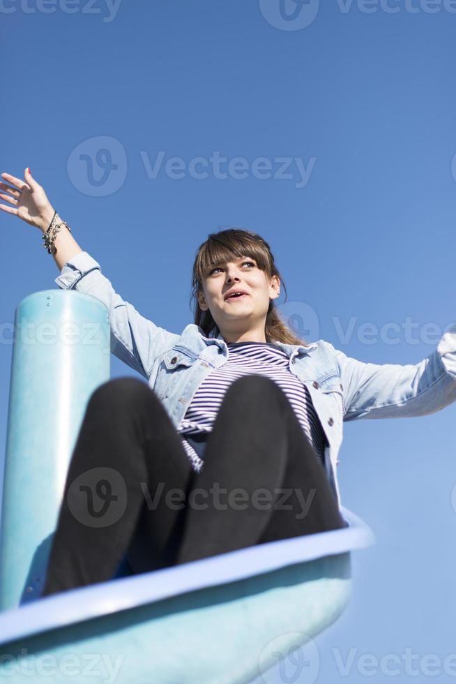 Eine infantile und glückliche Frau, die auf einer Kinderrutsche auf einem Spielplatz sitzt foto