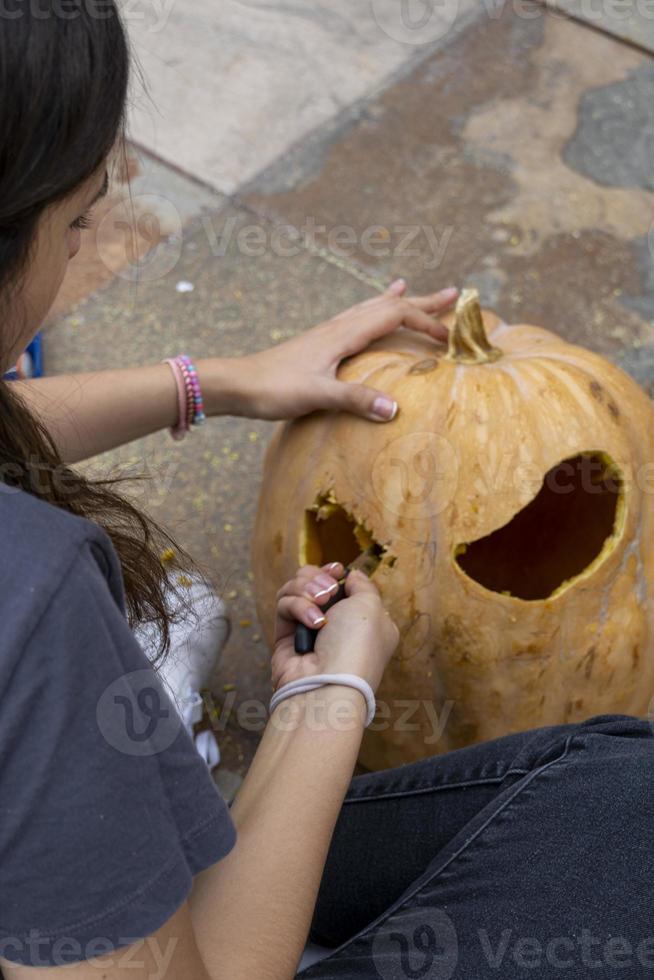 Frau, die großen orangefarbenen Kürbis für Halloween schnitzt, während sie zu Hause am Holztisch sitzt foto