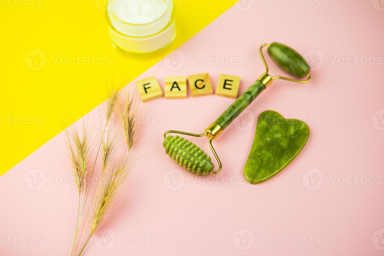 grüne gua sha gesichtsmassage werkzeuge. Rolle aus grüner Quarzjade auf rosa-gelbem Grund. Sahneglas, Aufschrift Gesicht in Holzbuchstaben. Nahaufnahme, Draufsicht. foto