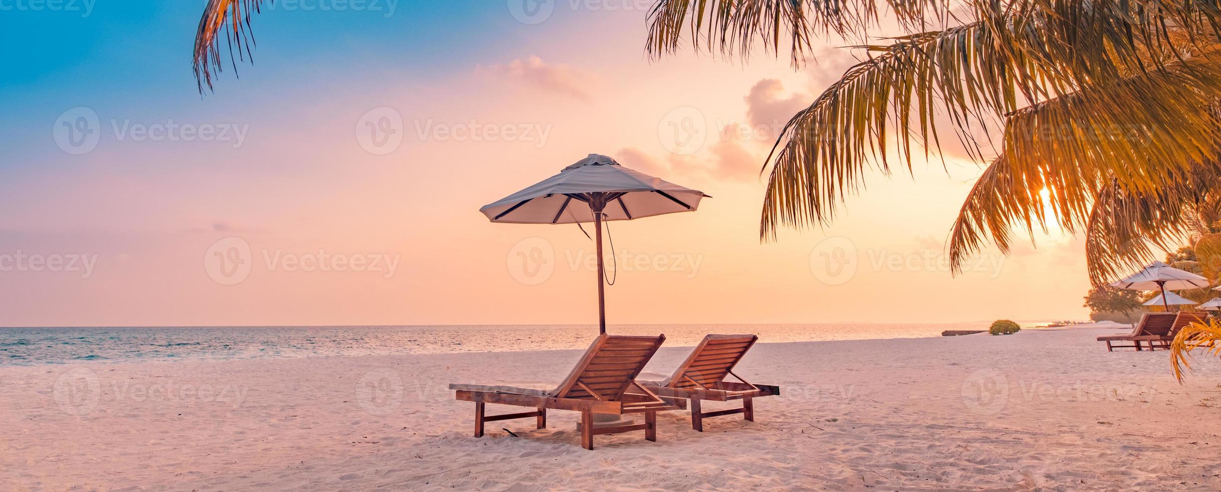 sommerliche Strandlandschaft. luxusurlaubs- und urlaubskonzept, sommerreisebanner. Panoramalandschaft mit Sonnenuntergangsstrand, zwei Liegestühlen, Sonnenschirm, Palmblatt, farbenfroher Sonnenuntergangshimmel für den Blick auf die paradiesische Insel foto