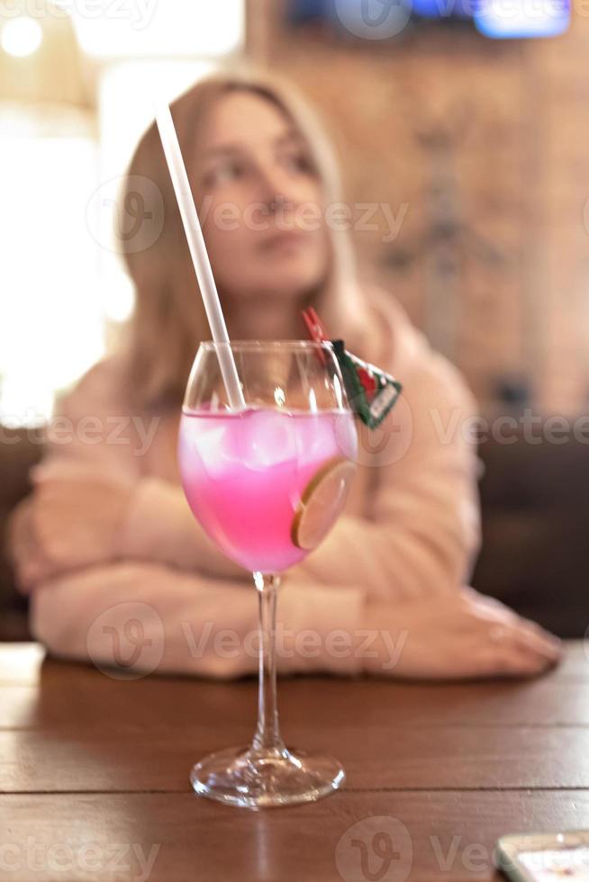 Fruchtiger rosafarbener Cocktail mit einer Limettenscheibe auf einem Tisch in einem Café. im Hintergrund eine verschwommene Gestalt einer Frau, die an einem Tisch sitzt foto