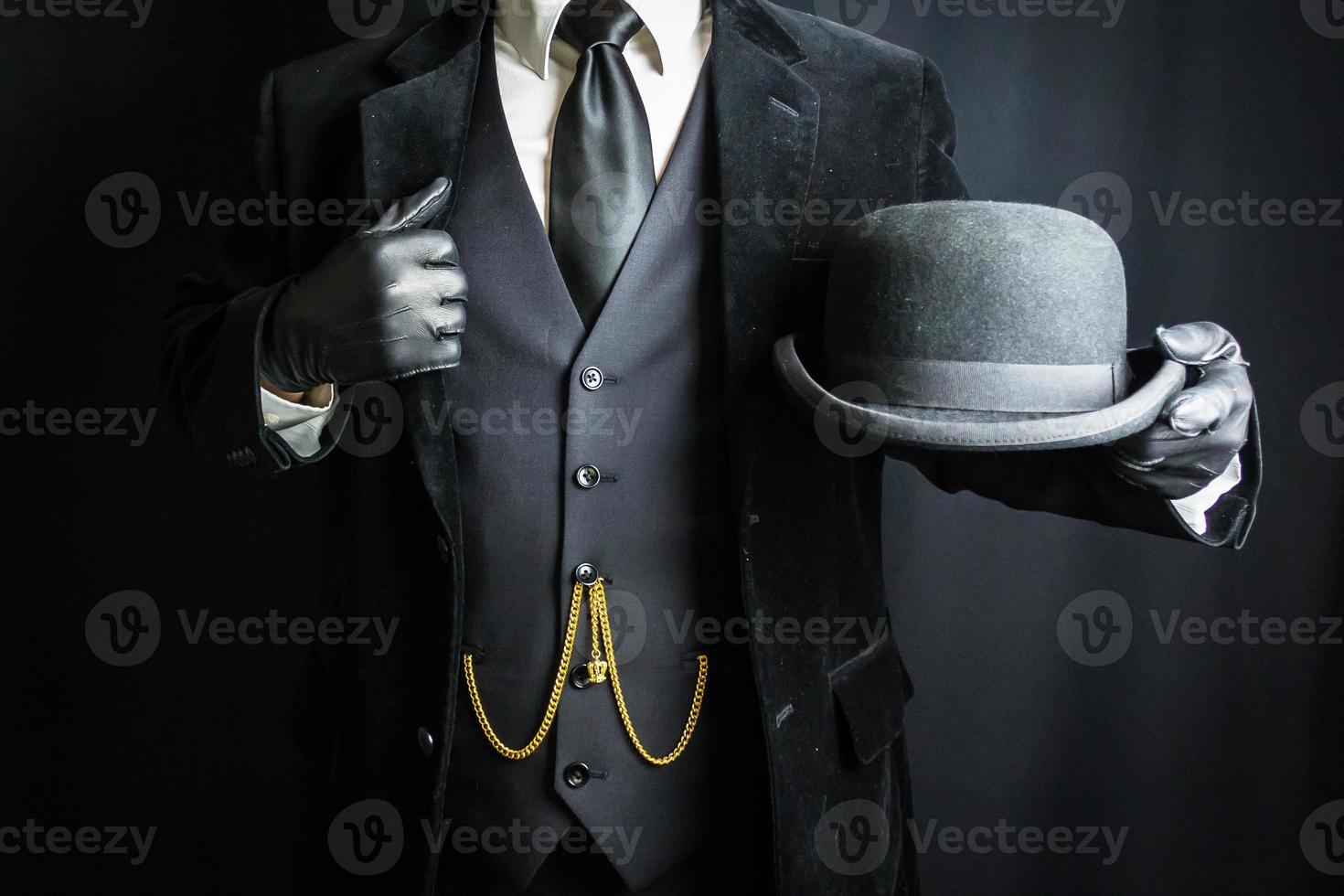 Mann in dunklem Anzug und Lederhandschuhen mit Melone auf schwarzem Hintergrund. Konzept des klassischen und exzentrischen britischen Gentleman foto