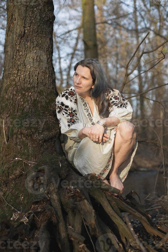 Nahaufnahme einer Frau im Leinenkleid mit Stickerei, die in der Nähe eines Baumporträtbildes sitzt foto