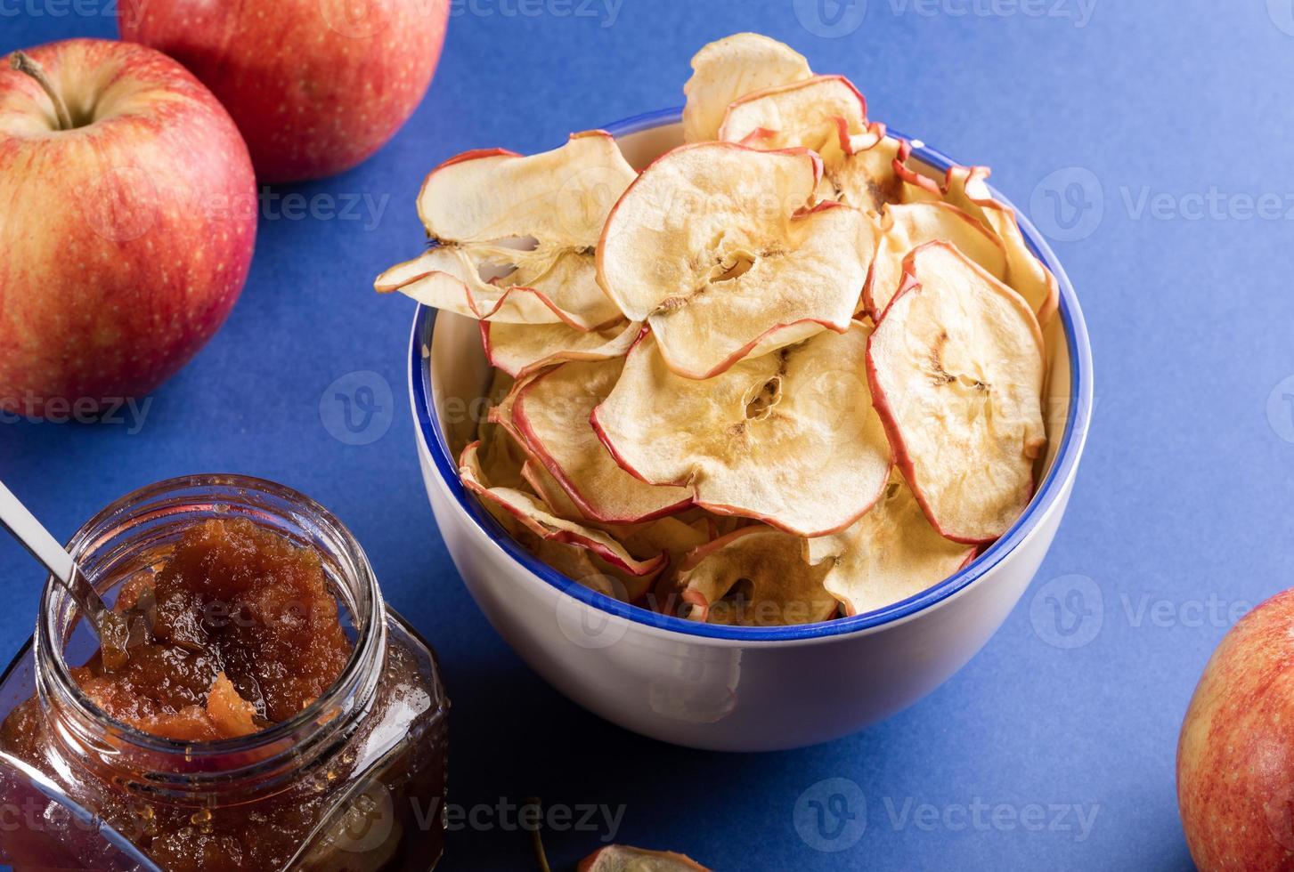 Weiße Keramiktasse mit Apfelchips, frischen roten Äpfeln und Glas mit Apfelmarmelade auf blauem Hintergrund. foto