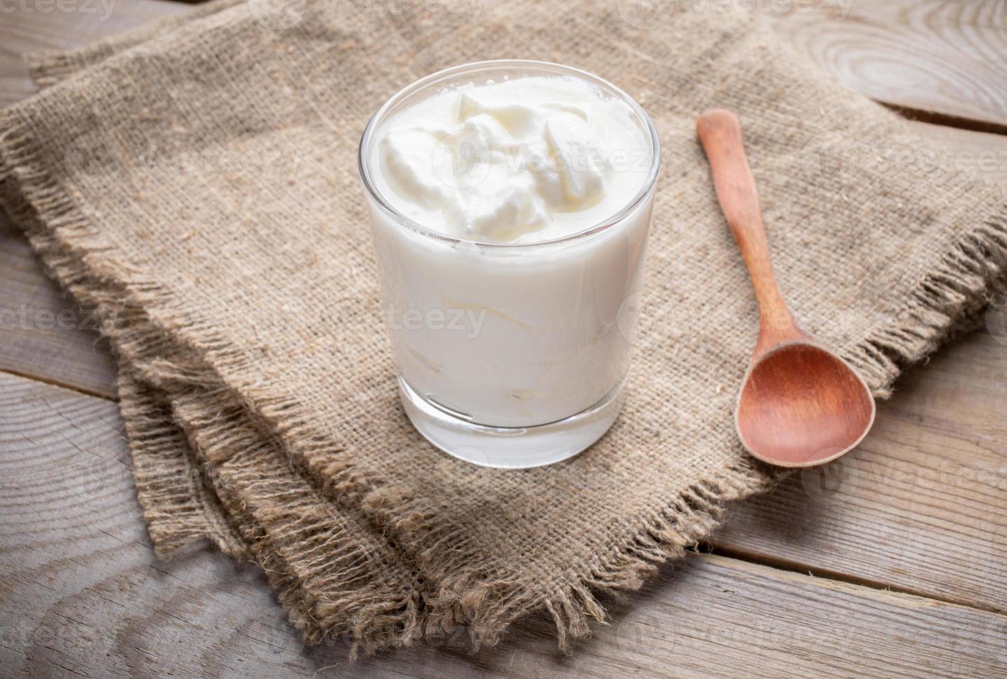 ein glas fetter hausgemachter joghurt mit holzlöffel steht auf sackleinenserviette auf holztisch. foto