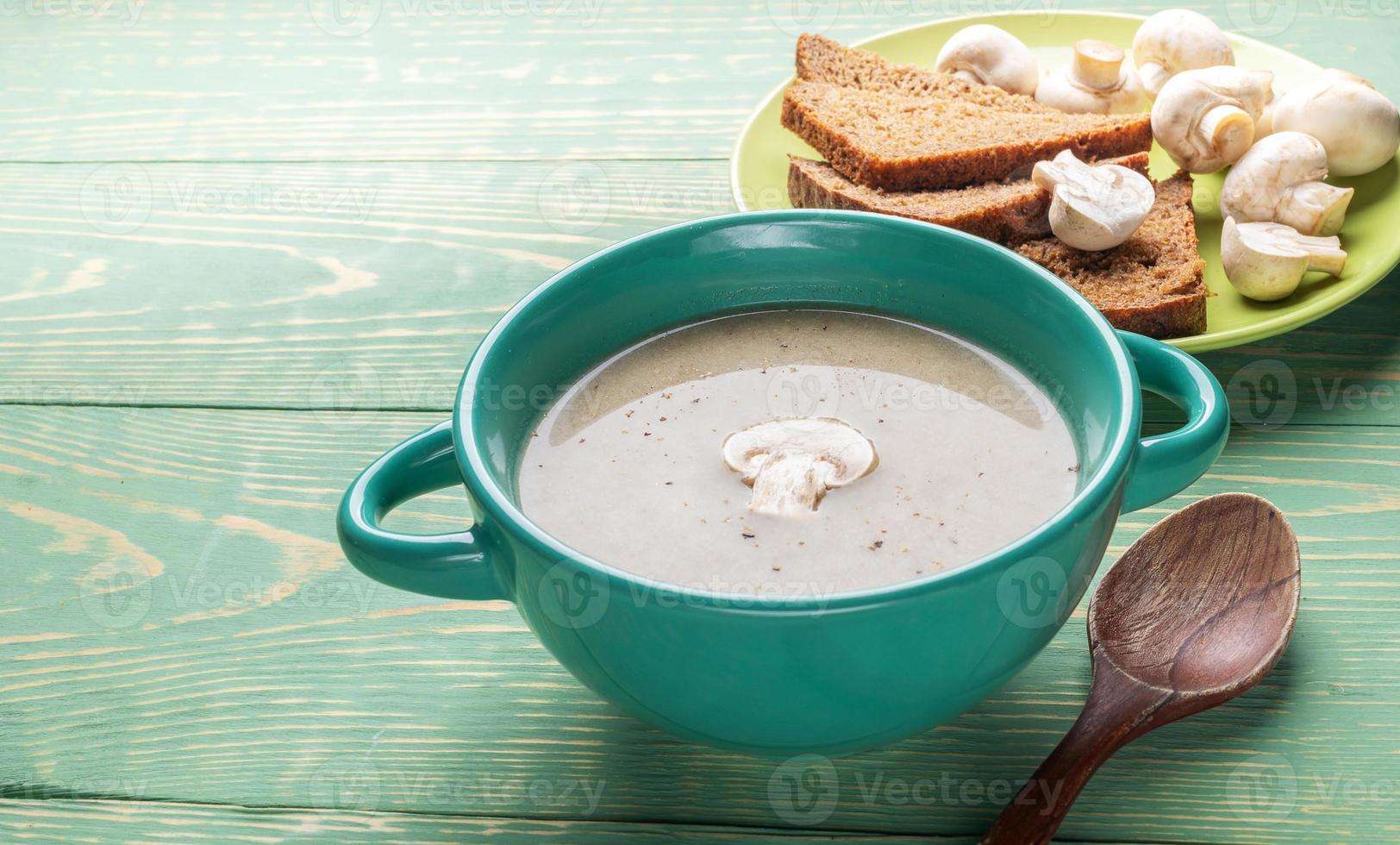 Türkisfarbene Keramikschale mit Pilzcremesuppe, Holzlöffel, Knoblauch, Brot, auf grünem Holzhintergrund. foto