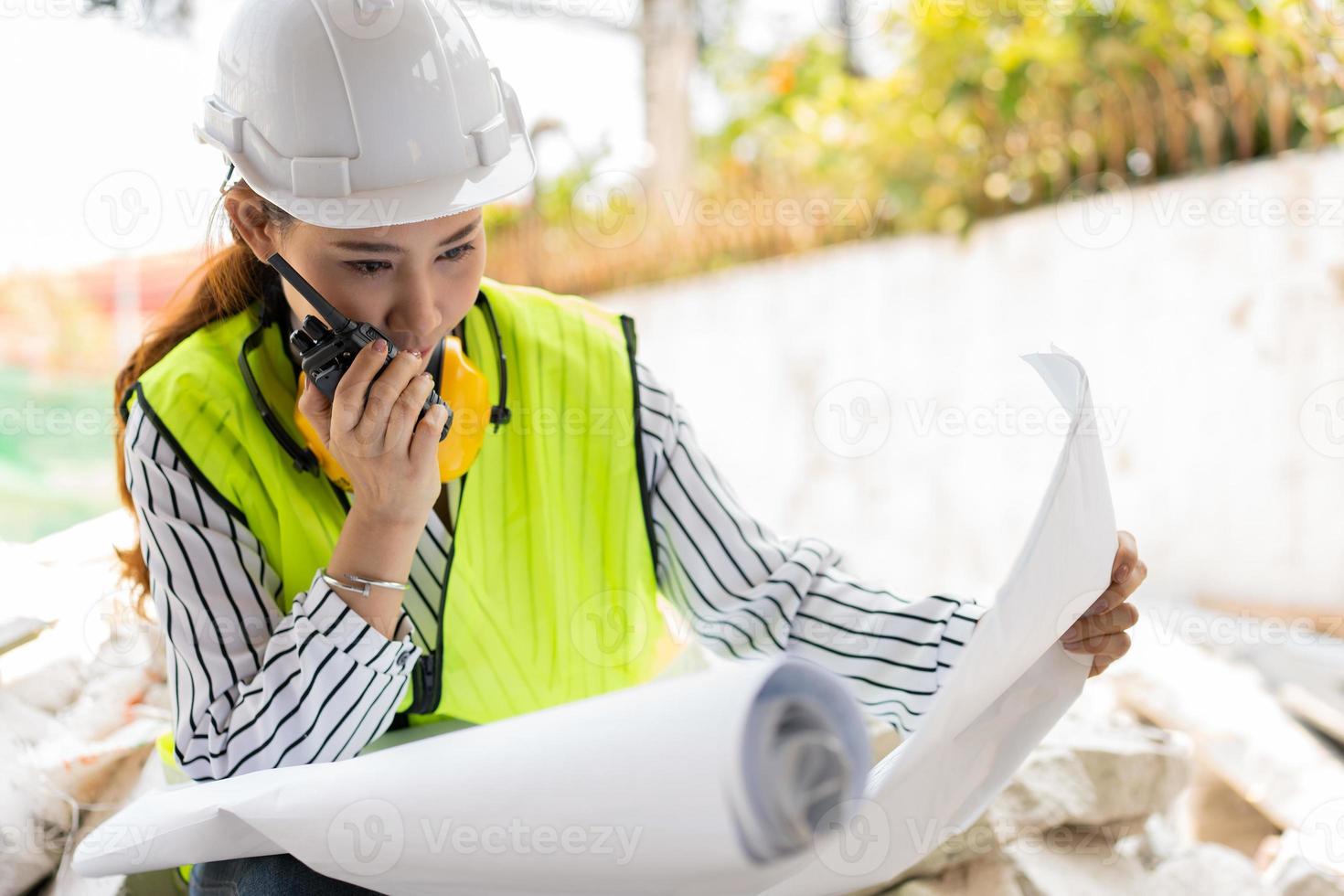 asiatische ingenieure oder junge architekten setzen aus sicherheitsgründen einen helm auf und sehen sich den bauplan an, um die baustelle der gebäudefabrik zu inspizieren, und verwenden das walkie-talkie für ein gespräch mit einem auftragnehmer. foto