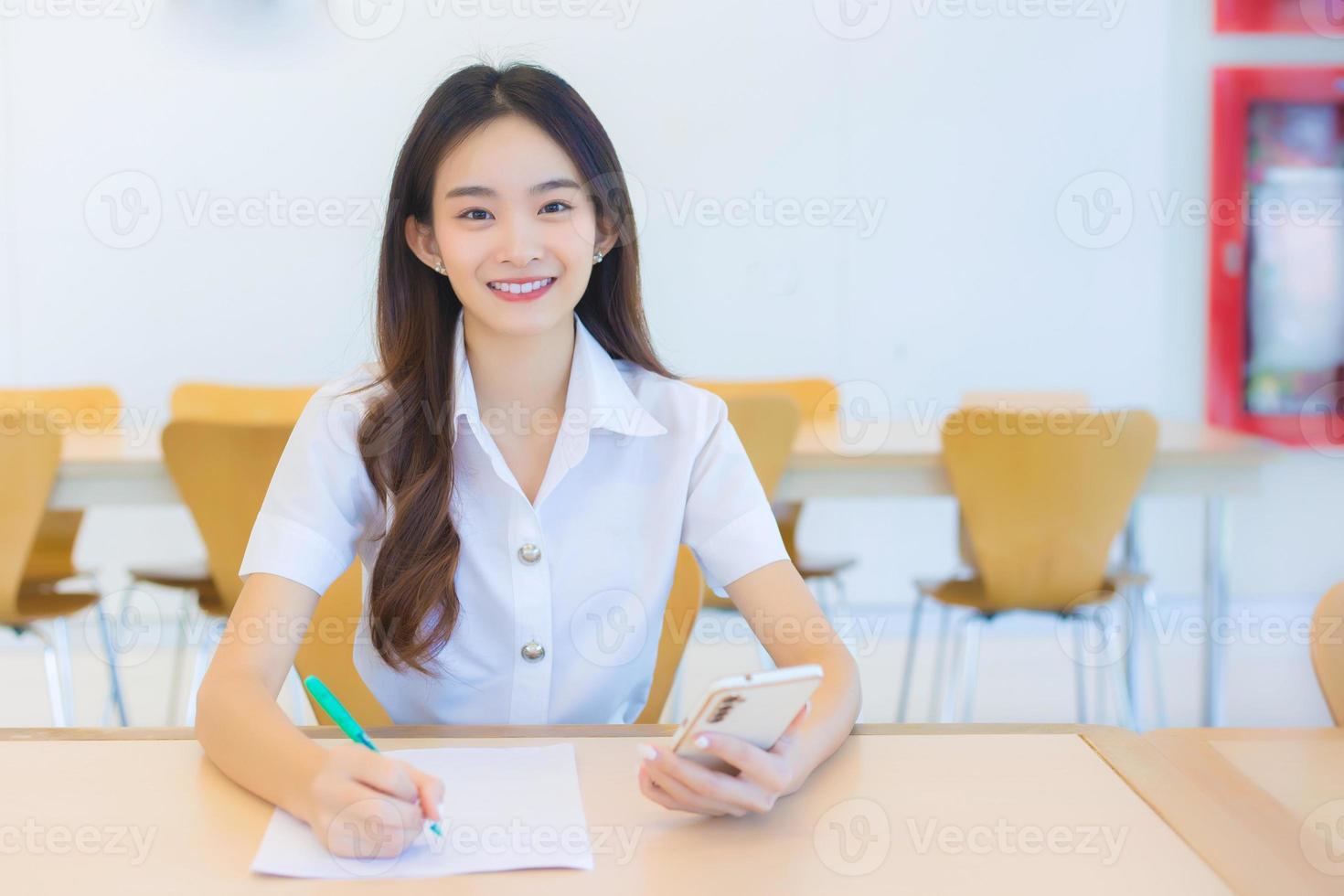 junge asiatische studentin in uniform, die smartphone benutzt und etwas über die arbeit schreibt. es gibt viele dokumente auf dem tisch, ihr gesicht mit einem lächeln in einem arbeiten, um informationen für einen studienbericht zu suchen foto