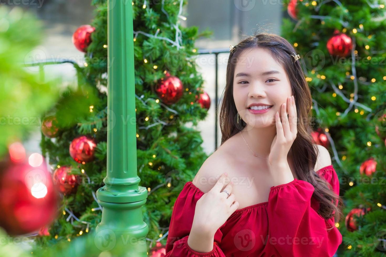 schöne asiatische frau im roten kleid steht glücklich lächelnd in die kamera vor dem weihnachtsbaum mit bokeh als hintergrund im thema weihnachts- und neujahrsfeiern foto
