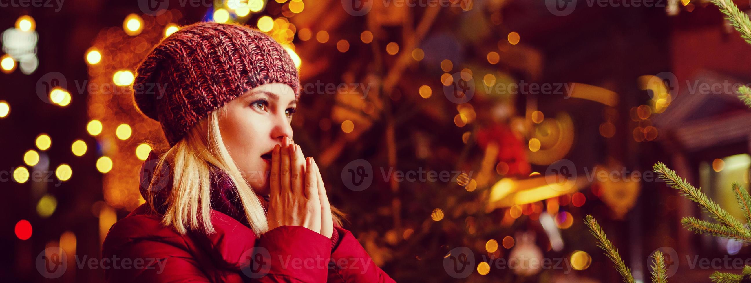 Outdoor-Foto von jungen schönen glücklichen lächelnden Mädchen, die auf der Straße posieren. festliche weihnachtsmesse im hintergrund. Modell mit stylischem Wintermantel, Strickmütze, Schal. foto