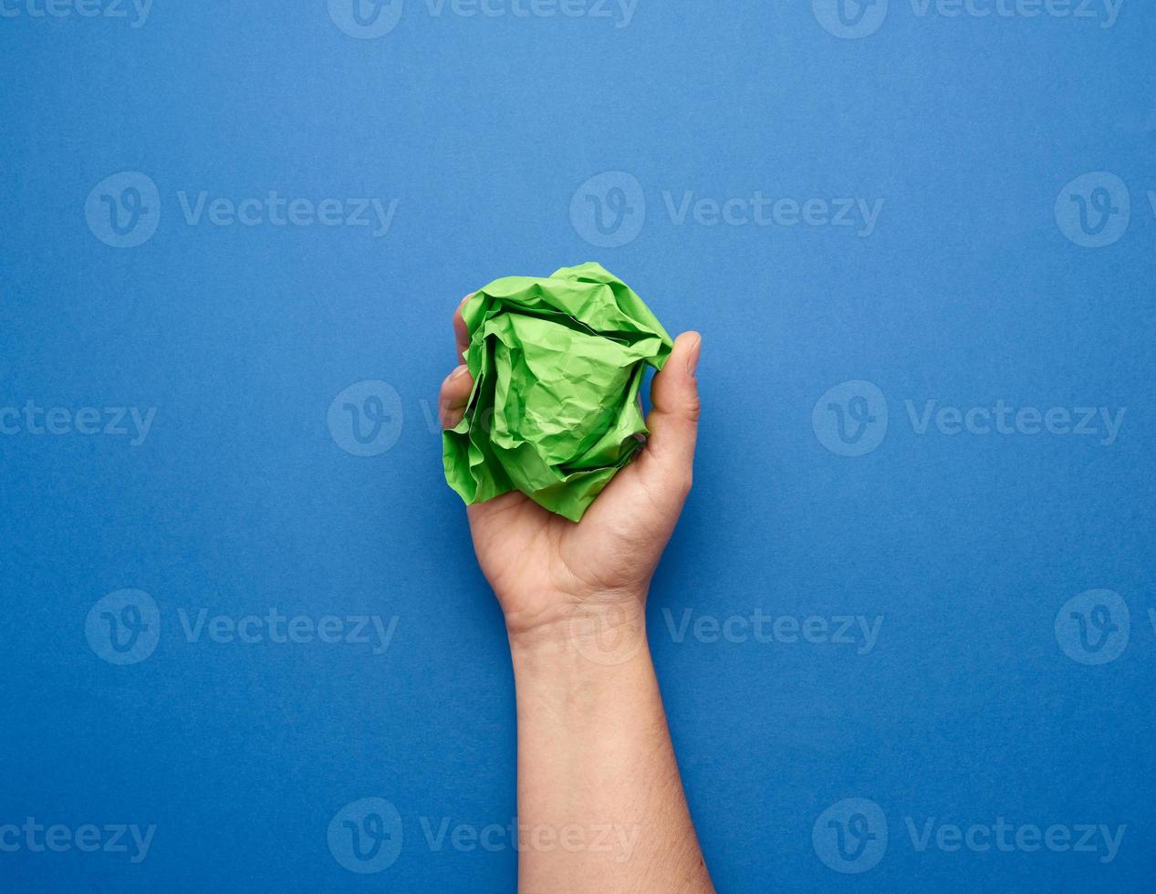 weibliche Hand, die einen zerknitterten Ball aus grünem Papier auf blauem Hintergrund hält foto