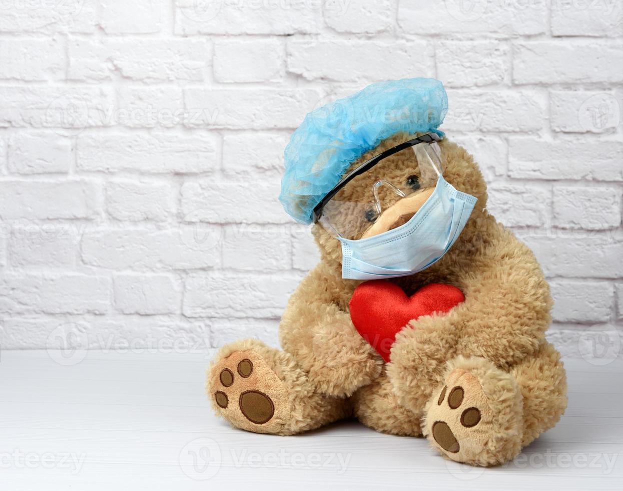 brauner teddybär sitzt in einer schützenden plastikbrille, einer medizinischen einwegmaske und einer blauen kappe foto