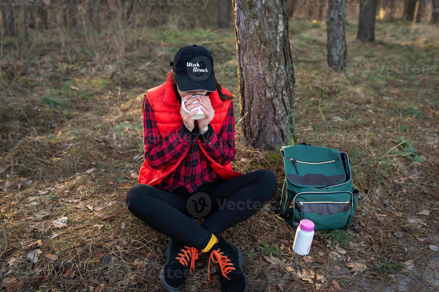 Trekking, Wandern. ein Rucksack mit Thermoskanne und Tasse steht neben einem Baum, ein Mädchen wärmt sich mit heißem Tee, einem Snack im Freien.Reisen und Tourismus foto