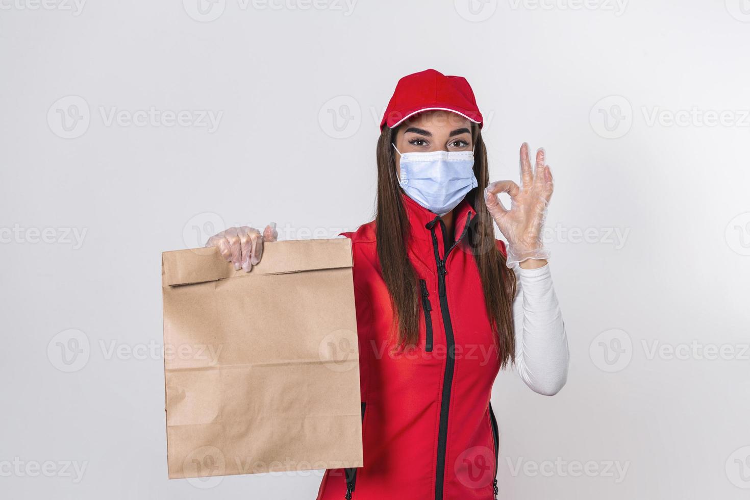 lieferfrau in roter uniform hält kraftpapierpaket mit lebensmitteln isoliert auf weißem hintergrund, trägt medizinische maske und handschuhe. weibliche angestellte ich arbeite als kurier. Service-Konzept. foto