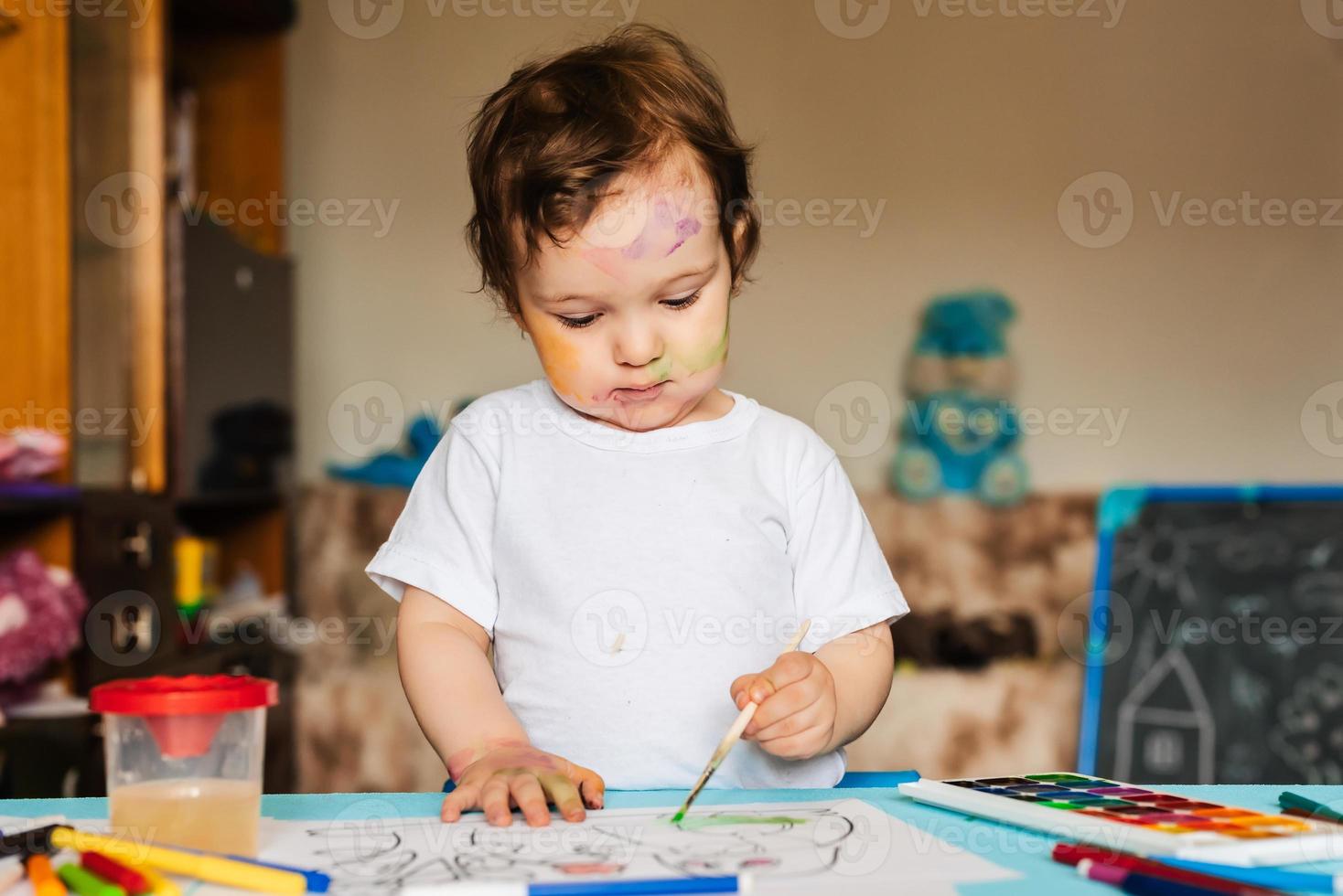 Fröhliches, fröhliches Kind, das mit Pinsel im Album zeichnet und dabei viele Malwerkzeuge verwendet. foto