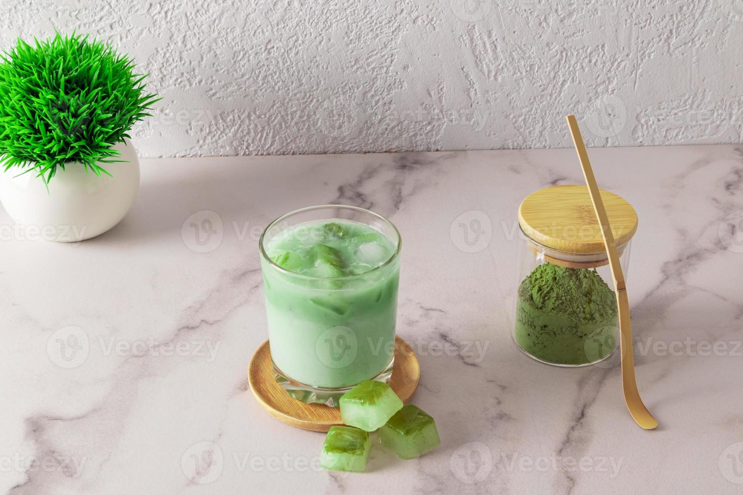 ein glas belebender kalter match latte mit eiswürfeln aus tee steht auf einer holzplatte. Matcha-Pulver in einem Glasgefäß. eine Alternative zu Kaffee. foto