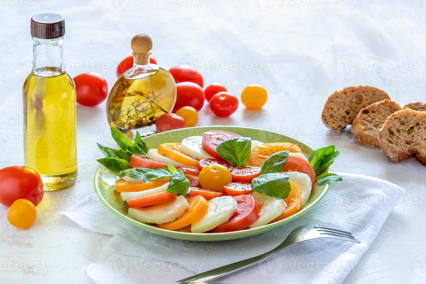 grüner teller mit traditionellem italienischem caprese-salat und olivenölflaschen, tomaten und brot auf weißem tisch. foto