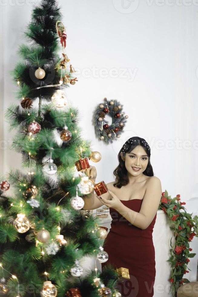 Porträt eines hübschen jungen Mädchens, das den Weihnachtsbaum schmückt und lächelnd ein rotes Kleid im geschmückten Weihnachtswohnzimmer im Innenbereich trägt foto