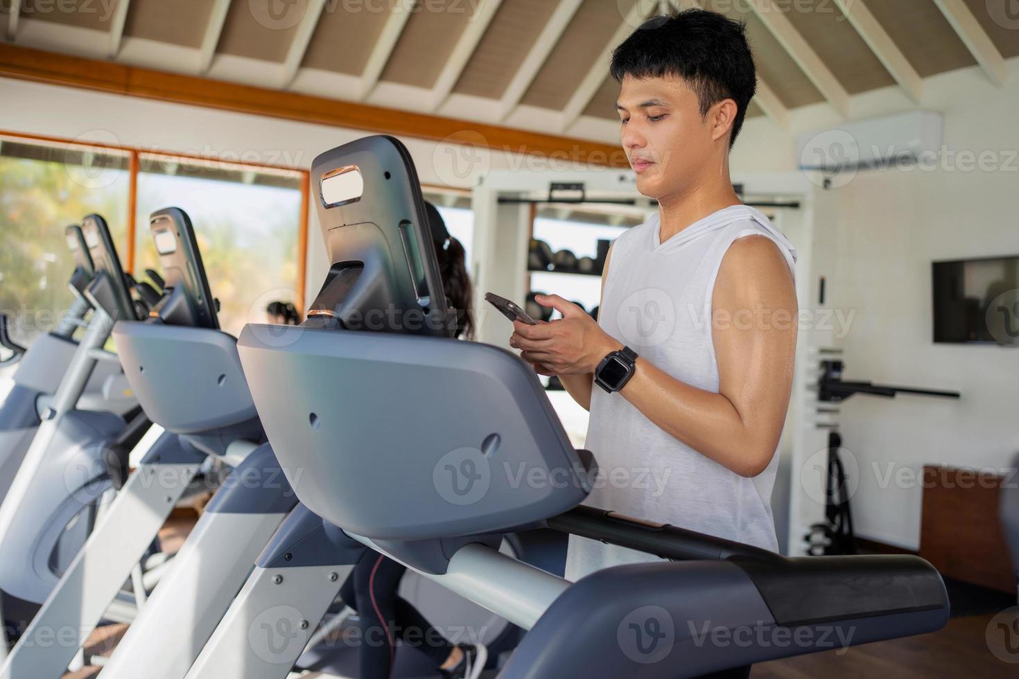 junger asiatischer mann, der smartphone auf laufband im fitnesscenter verwendet. foto