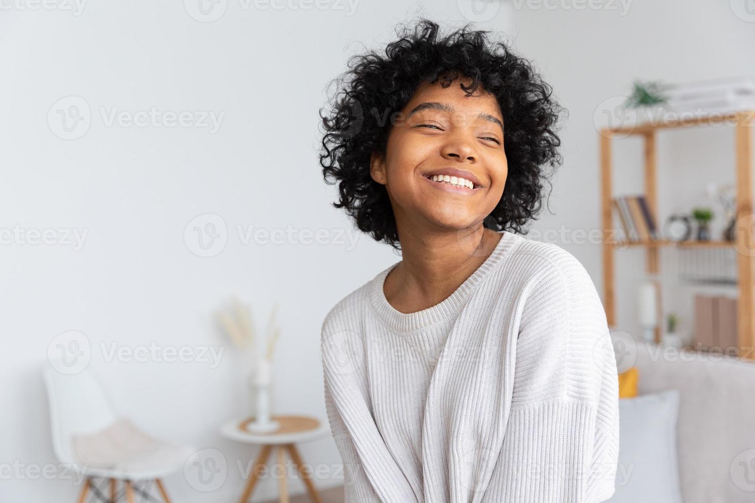 schönes afroamerikanisches mädchen mit afrofrisur, das zu hause drinnen lächelt. junge afrikanische frau mit lockigem haar, die im wohnzimmer lacht. freiheit glück sorglos glückliche menschen konzept. foto
