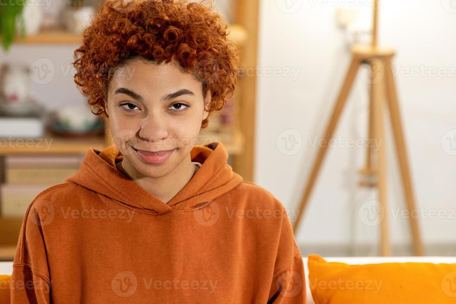 Schönes afrikanisch-amerikanisches Mädchen mit Afro-Frisur lächelnd auf dem Sofa zu Hause drinnen sitzend. Junge afrikanische Frau mit lockigem Haar lächelt. freiheit glück sorglos glückliche menschen konzept. foto