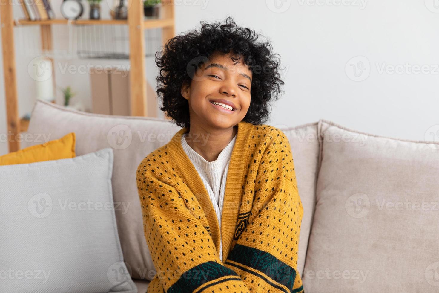 Schönes afrikanisch-amerikanisches Mädchen mit Afro-Frisur lächelnd auf dem Sofa zu Hause drinnen sitzend. Junge afrikanische Frau mit lockigem Haar lacht. freiheit glück sorglos glückliche menschen konzept. foto