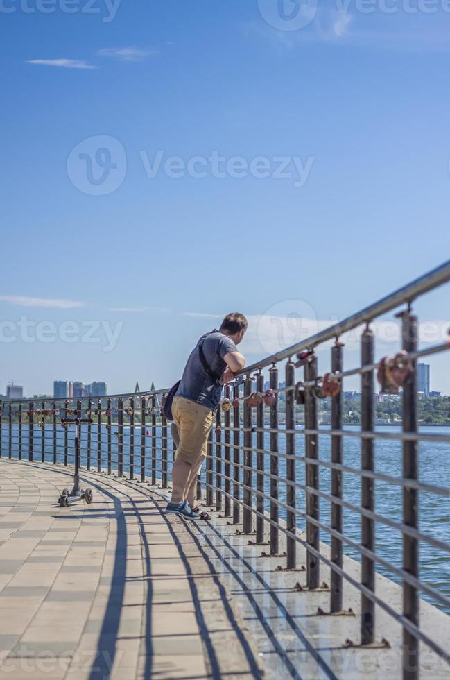Kinder und ein Mann gehen über die Brücke und schauen auf den Fluss. Am Geländer sind verschiedene interessante Dinge befestigt. ein heißer Sommertag foto