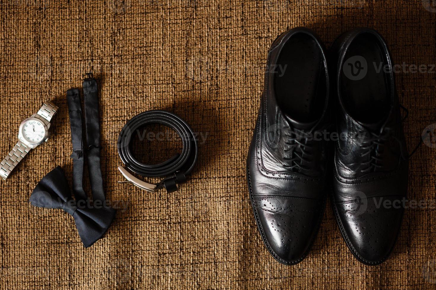 Schwarze Schuhe, schwarzer Gürtel, schwarze Uhr, schwarzer Schmetterling, Manschettenknöpfe und Parfüm auf braunem Hintergrund mit Sackleinen foto