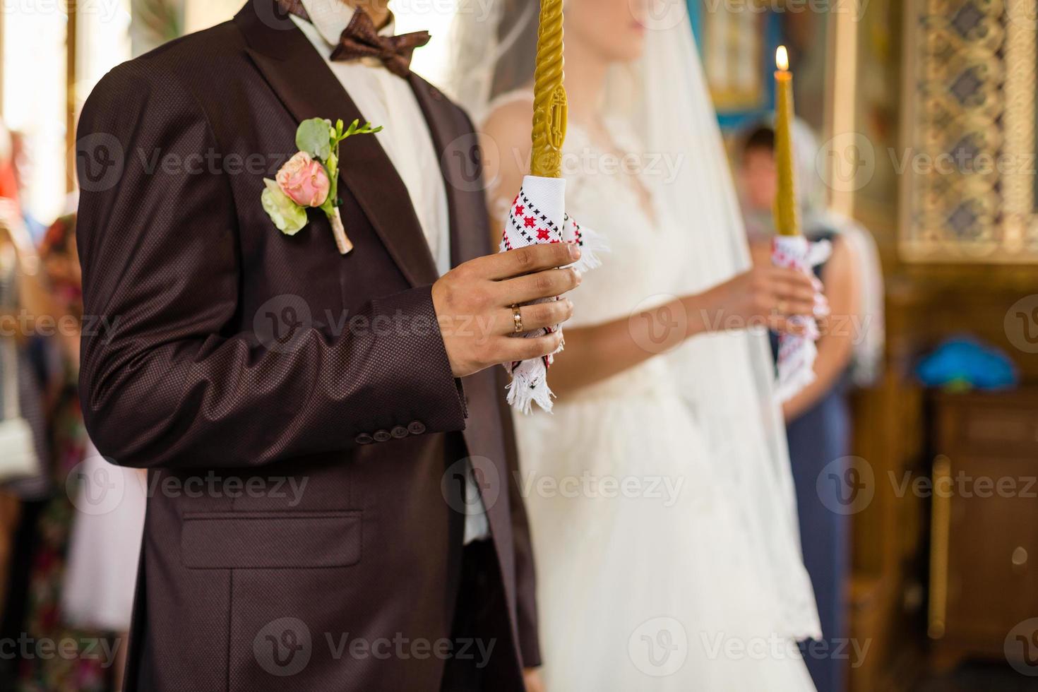 wunderschöne braut und stilvoller bräutigam, die kerzen bei der offiziellen hochzeitszeremonie in der alten kirche halten. traditioneller Augenblick foto