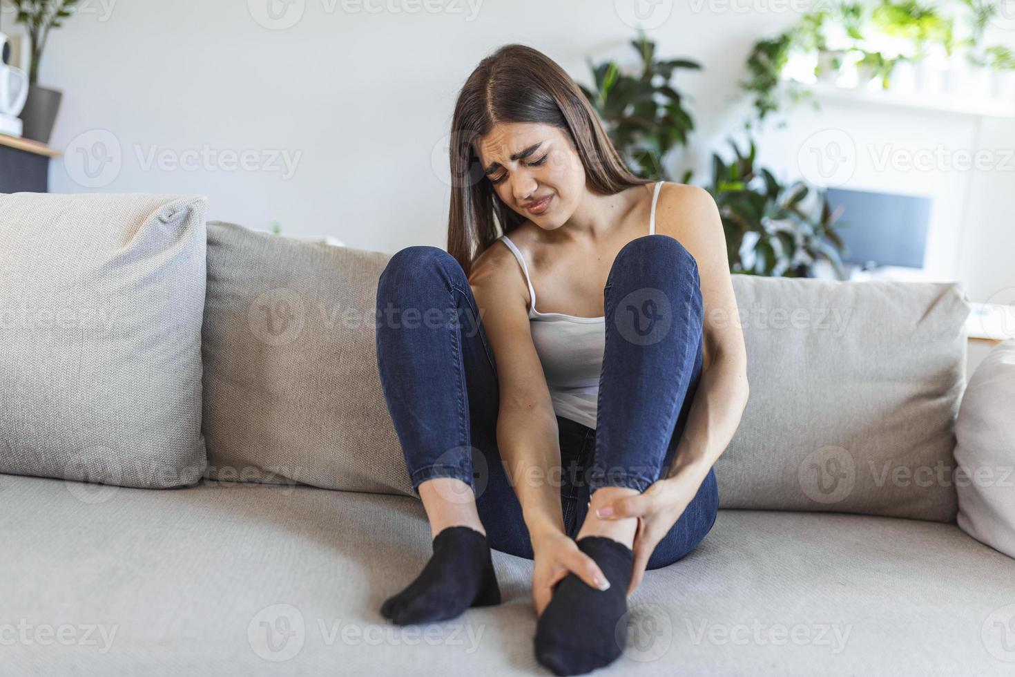 Hände massieren geschwollene Füße, während sie tagsüber zu Hause auf dem Sofa sitzen. Foto einer jungen kaukasischen Frau, die unter Schmerzen im Bein leidet. Frau, die ihre Beine nach dem ganzen Tag bei der Arbeit im Büro massiert