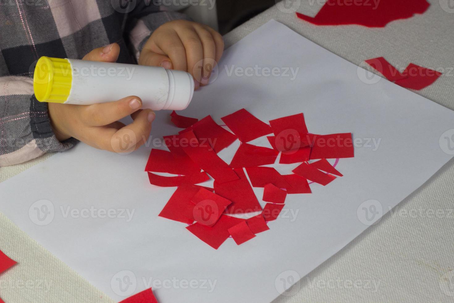 Kleines Mädchen, das rote Herzapplikationen mit Papier und Kleber auf weißem Blatt macht, Valentinstagsgrußkarte, Weihnachtsgeschenk für Muttertag, handgemachtes Kindergeschenk, Liebessymbol mit Papier, Kleinkindaktivität foto