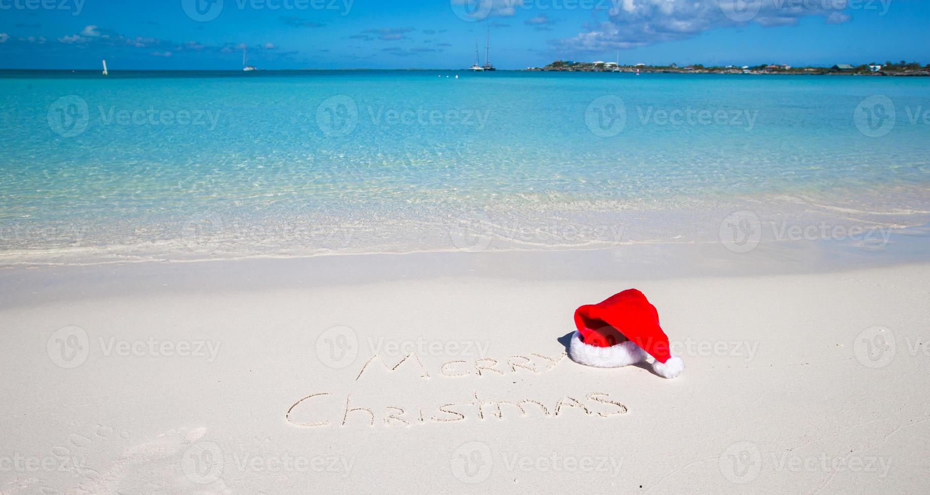 weihnachtsmütze am weißen sandstrand und frohe weihnachten in den sand geschrieben foto