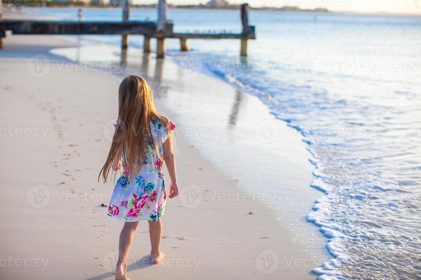 Entzückendes kleines Mädchen, das bei Sonnenuntergang am weißen tropischen Strand spazieren geht foto