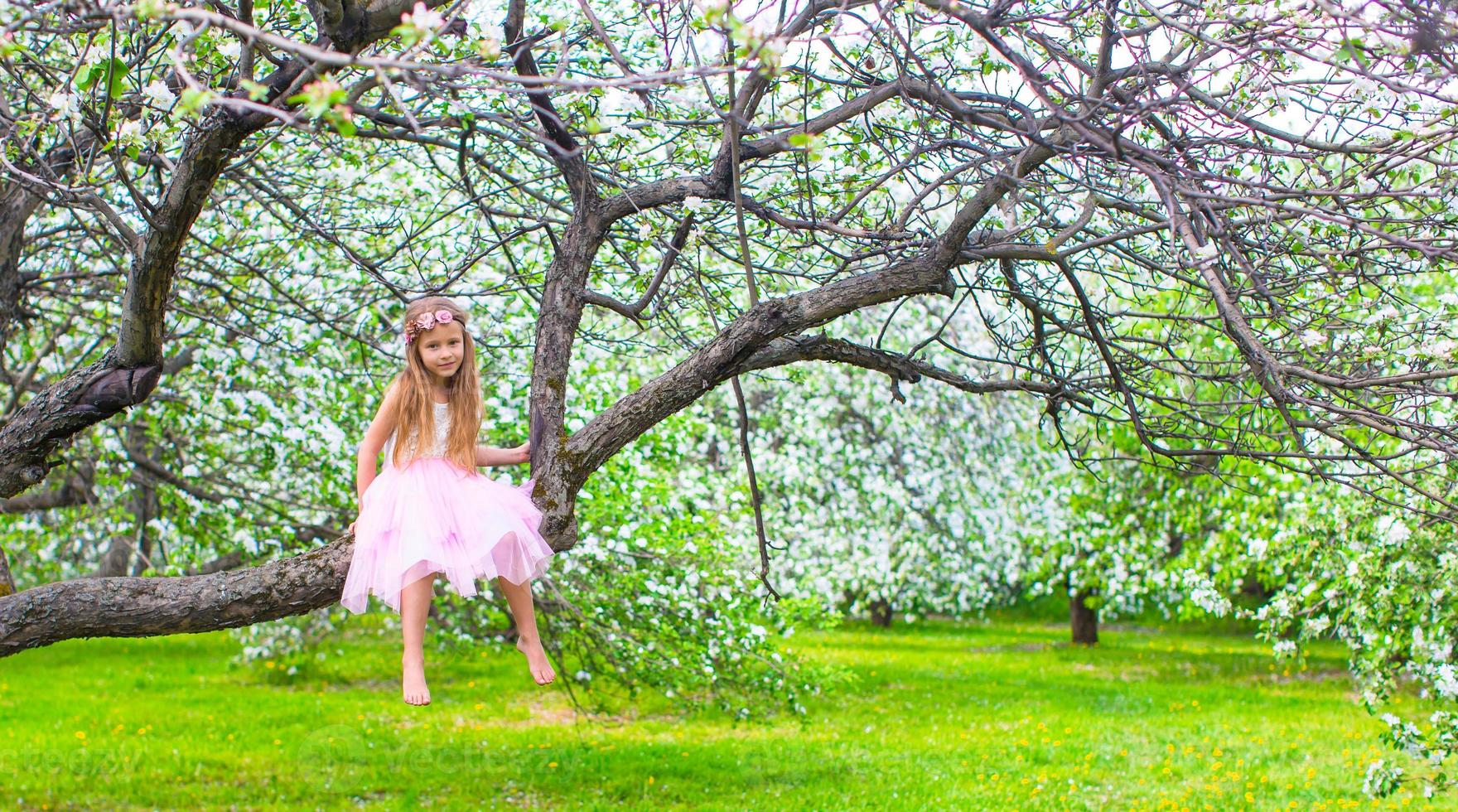 Kleines entzückendes Mädchen, das auf blühendem Baum im Apfelgarten sitzt foto