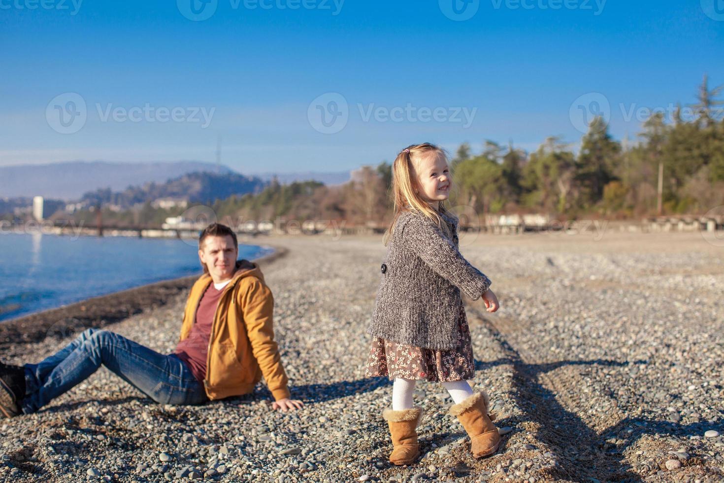 Entzückendes kleines Mädchen mit Vater, das sich am warmen Wintertag am Strand amüsiert foto