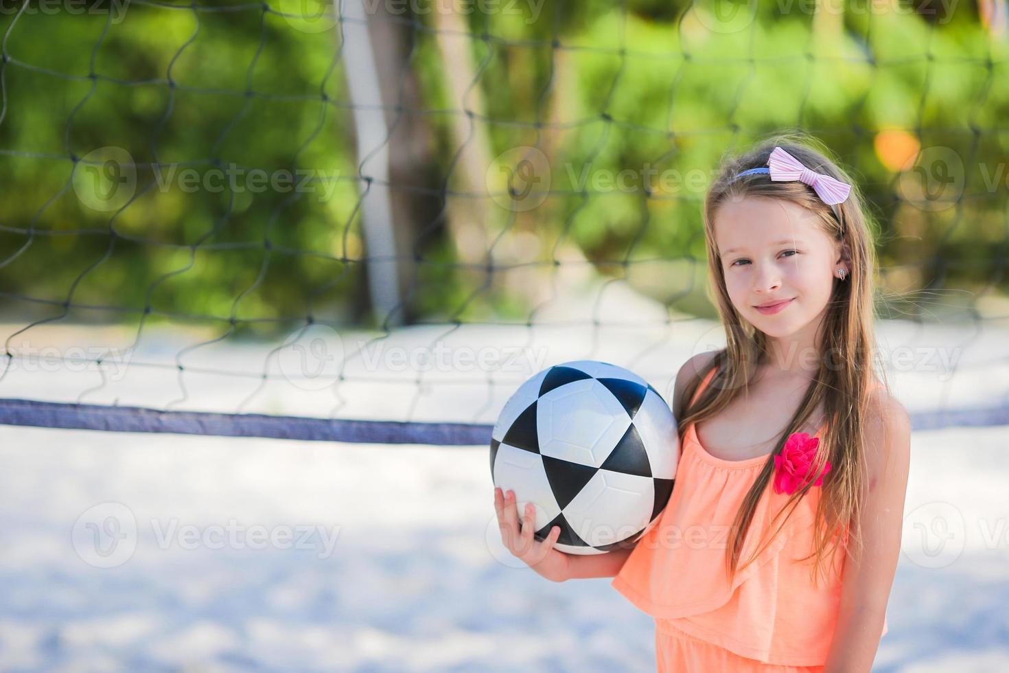 Kleines entzückendes Mädchen, das Volleyball am Strand mit Ball spielt foto