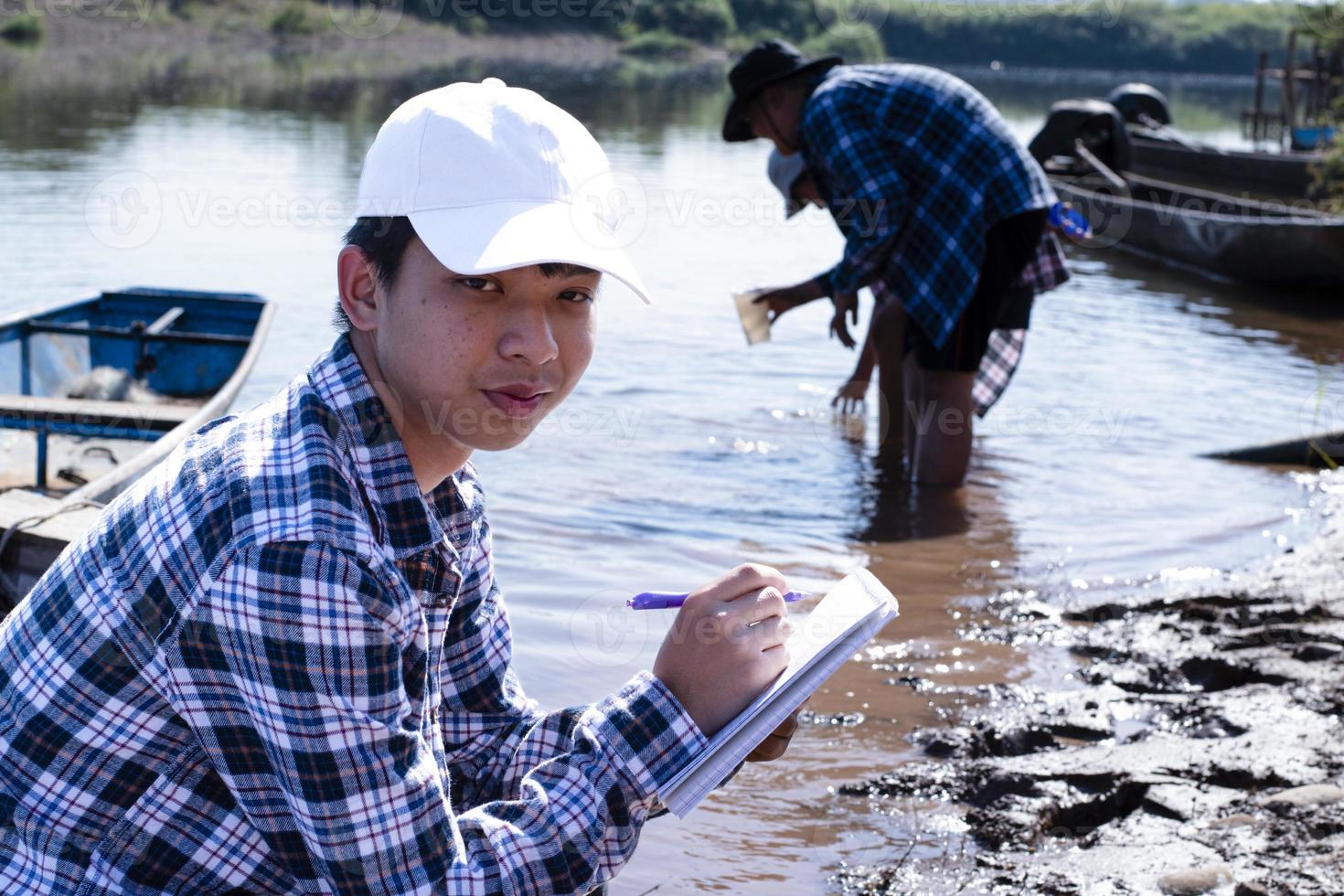 Der junge asiatische Junge hält ein transparentes Rohr, in dem sich Beispielwasser befindet, um das Experiment und die pH-Wert-Messung durchzuführen, während sein Schulprojekt mit seinen Freunden hinter dem Fluss arbeitet, an dem er lebte. foto