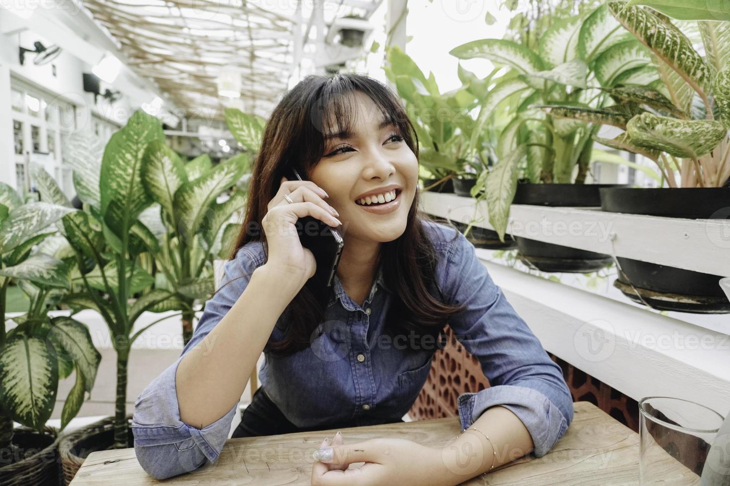 eine glückliche asiatische frau in einem restaurant, die ein blaues hemd trägt und ihr telefon hält foto