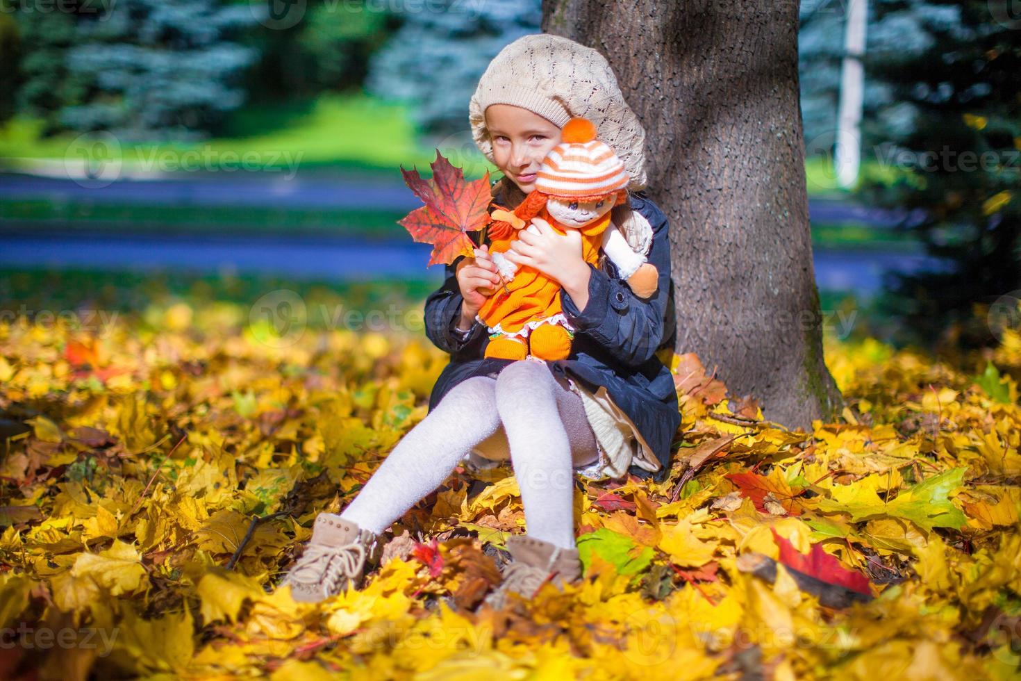 hübsches modemädchen, das an einem sonnigen herbsttag unter einem ahornbaum mit puppe und rotem blatt sitzt foto