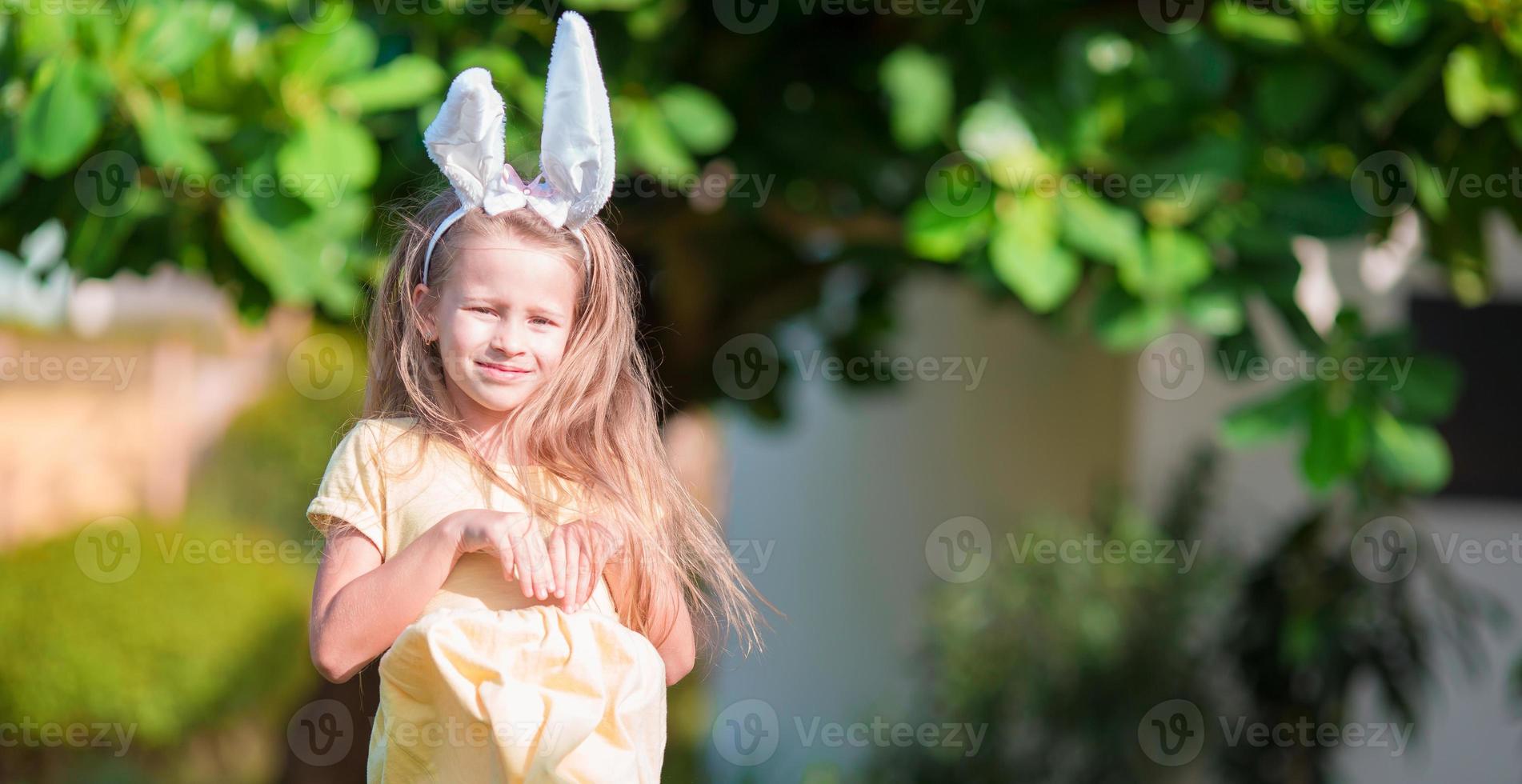 Entzückendes kleines Mädchen, das am Frühlingstag Hasenohren mit Ostereiern trägt foto