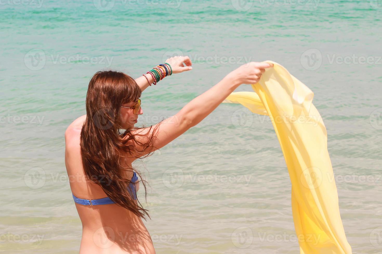 Schönes junges Mädchen, das im Strandurlaub gelben Stoff bei Wind hält foto