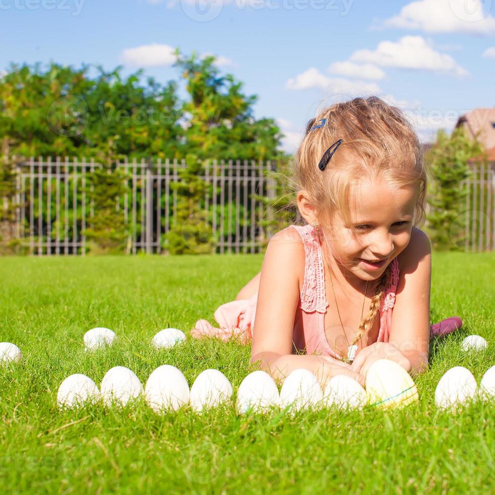 Porträt eines kleinen glücklichen Mädchens, das mit weißen Ostereiern auf grünem Gras spielt foto