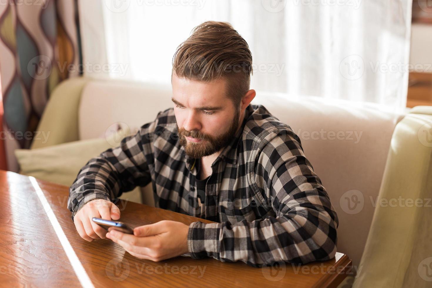 junger Hipster-Typ, der Nachrichten aus sozialen Netzwerken sucht, die in der Freizeit im Café per Smartphone gelesen werden foto