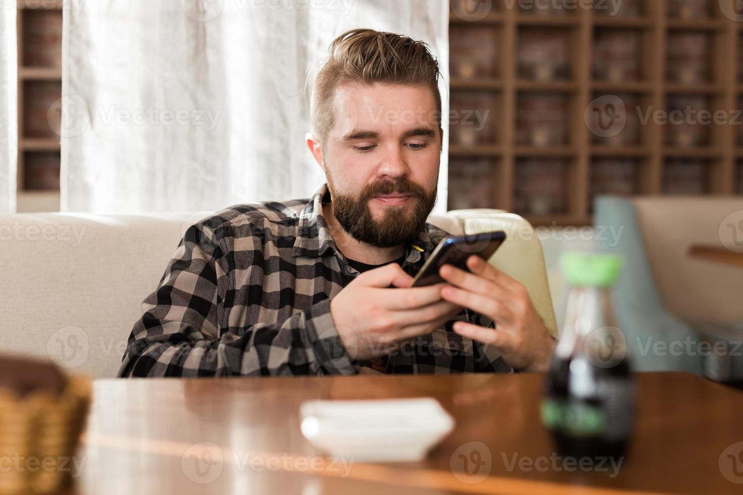 junger Hipster-Typ, der Nachrichten aus sozialen Netzwerken sucht, die in der Freizeit im Café per Smartphone gelesen werden foto