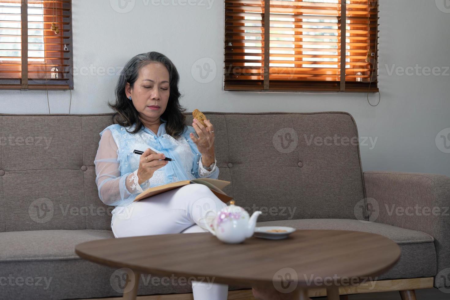 Porträt, wunderschöne Asiatin im Alter von 60 Jahren in Freizeitkleidung, die Kekse isst und ihr Tagebuch schreibt, während sie sich in ihrem Wohnzimmer entspannt foto