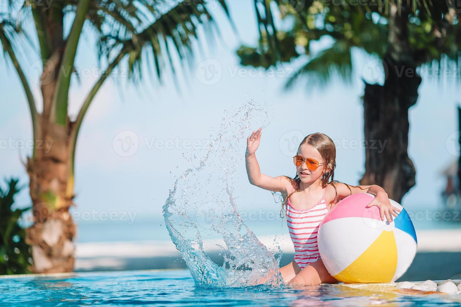 Lächelndes entzückendes Mädchen, das mit aufblasbarem Spielzeugball im Außenpool spielt foto