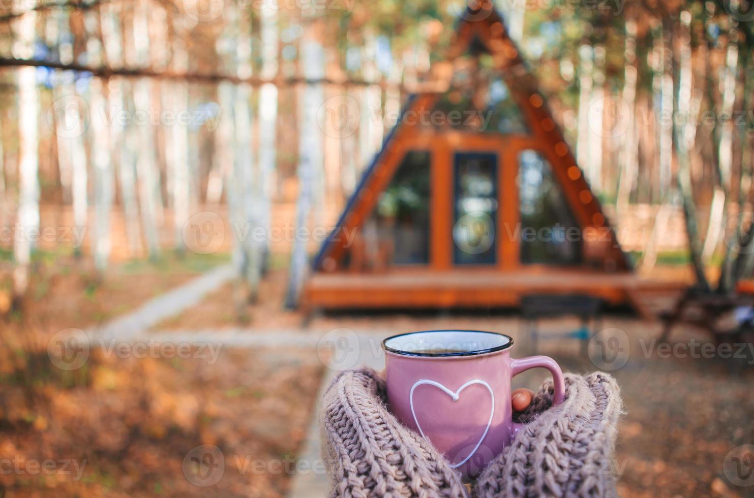 Heiße Tasse Tee, die die Hände der Frau im Wollpullover auf dem Hintergrund des gemütlichen Hauses wärmt foto