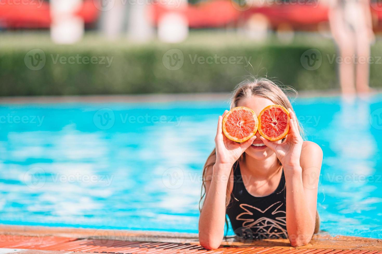 kleines Mädchen, das die Augen mit orangefarbenen Hälften in der Nähe der Augen auf dem Hintergrund-Swimmingpool bedeckt foto