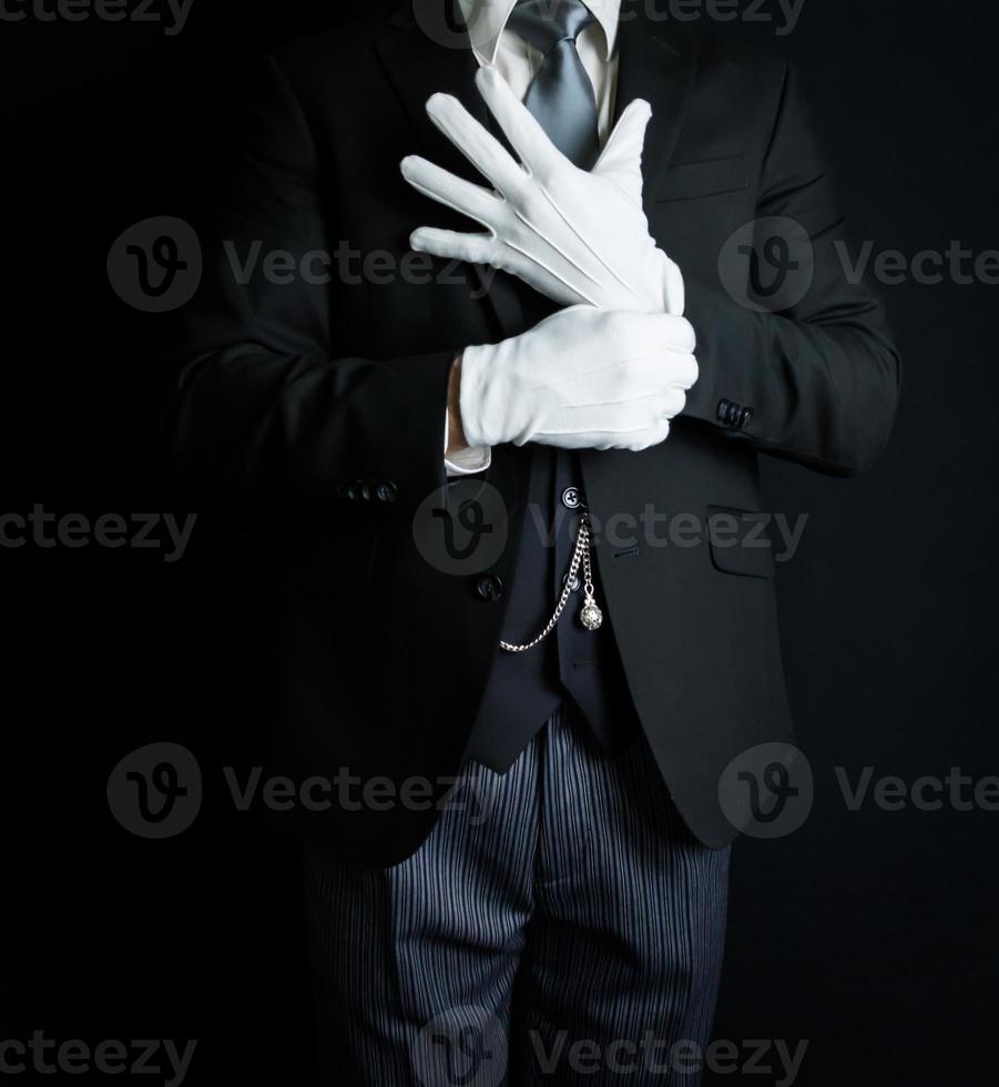 Porträt des Butlers im dunklen Anzug auf schwarzem Hintergrund, der saubere weiße Handschuhe anzieht. konzept der dienstleistungsbranche und der professionellen gastfreundschaft. foto